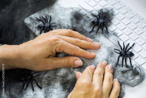 Hands working on a laptop surrounded by black spiders and cobweb decorations, creating a spooky Halloween atmosphere and concept of fear, digital threats and working from home.