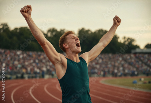 Young male athlete raises both fists and shouts in celebration on a red running track, with a blurred outdoor stadium crowd and trees behind him.