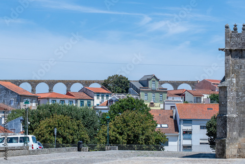 Historic stone aqueduct and red tiled roofs of an ancient town framed by carved granite finials under a clear blue sky.