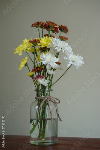 Colorful chrysanthemum flower bouquet in a clear glass bottle with a rustic twine bow, featuring white, yellow, and deep burgundy blooms on a dark wooden table against a neutral background