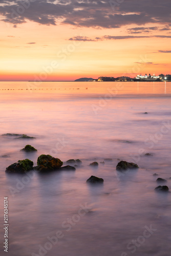Serene sunset over calm sea with rocks in foreground and distant city skyline