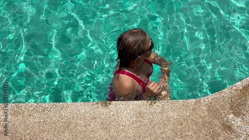 Woman swims in clear pool, making waves as they move. The sun shines brightly on water, reflecting light and creating a playful look. It is a warm day.