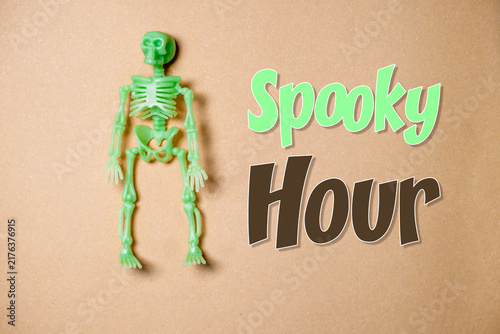 Картината върху платно Glow in the dark skeleton toy next to the phrase 'Spooky Hour' on a brown background, suggesting a Halloween theme