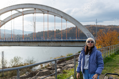 Cuadro en lienzo Adult woman wearing sunglasses, knitted beanie and blue puffer jacket stands beside Jezioro Mucharskie with the distinctive concrete arched bridge behind her