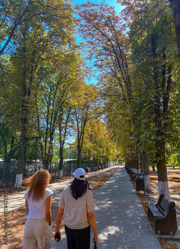 Two women seen from behind walking along a tree-lined park avenue