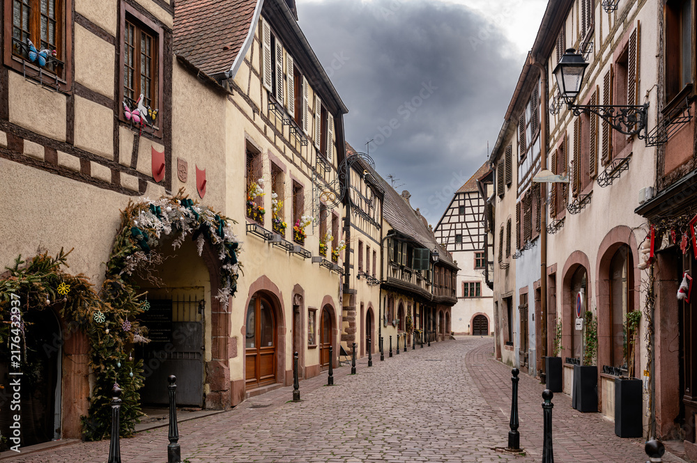 Fototapeta premium Thick garland of greenery and hanging ornaments arches over a doorway on a Kaysersberg street where sandstone arcades and shuttered facades run away under a heavy grey sky.