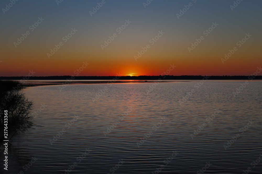 Obraz premium Beautiful sun setting behind forest horizon close up. Crimson orange sky twilight reflection on calm river water surface. Horizontal outdoor nature camping and fishing concept with copy space