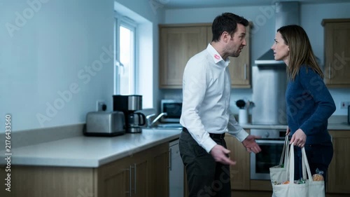 Woman confronting husband about lipstick mark on his collar in kitchen. Cheating spouse caught in act. Broken marriage, betrayal and domestic argument concept.