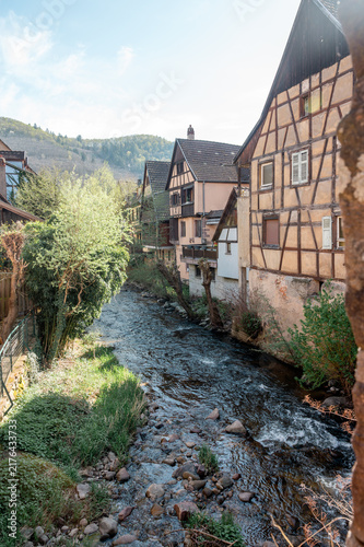 Obraz na płótnie Shallow rocky channel of the Weiss river runs between an ochre half timbered wall and a green bank in Kaysersberg with the wooded valley opening out under hazy spring light