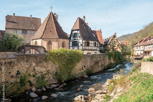 Quadro su tela Stone chapel with a steep tiled roof stands behind a long rampart wall above the Weiss river in Kaysersberg while half timbered houses step away toward the wooded valley side