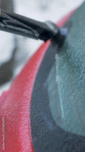 Male hand lifts rear wiper on red vehicle and removes brittle ice crust from blade. Frozen residue sticks to rubber, weakens contact with glass. Severe cold, icy street create hazardous conditions