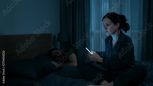 Woman checking smartphone in bed while husband sleeps. Secret messaging and betrayal causing marital crisis. Distant couple in bedroom at night, loneliness concept.