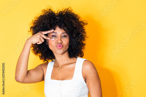 фотографія young latin woman making a peace sign gesture over her eye, puckering her lips p