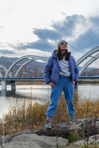 Fotografía Full-length portrait of an adult woman standing confidently on shoreline rocks overlooking Jezioro Mucharskie and its monumental arched bridge
