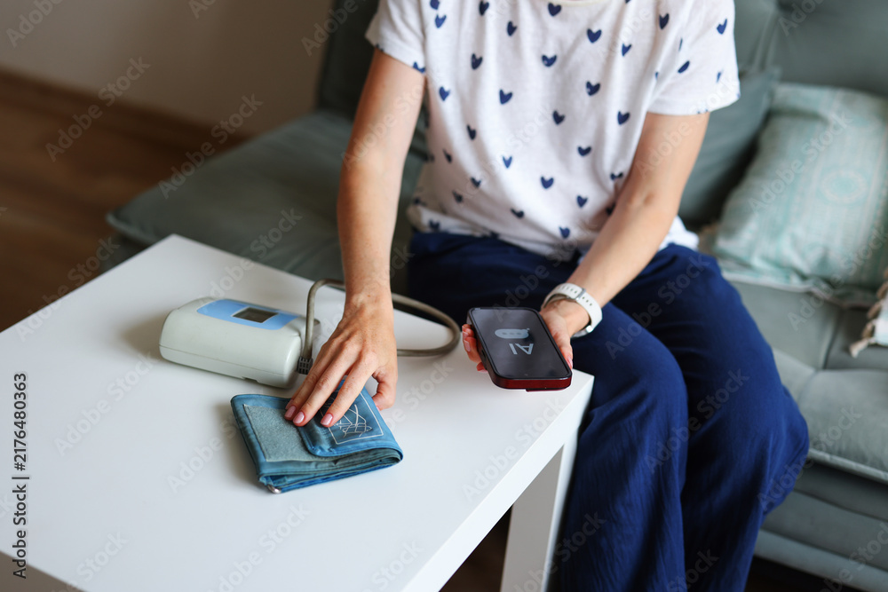 Fototapeta premium Woman chooses AI chatbot instead of doctor consultation for health concerns, using smartphone while measuring blood pressure at home.