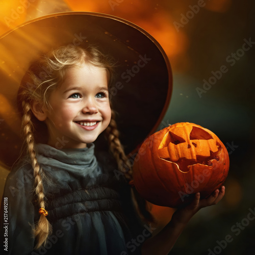 Cute little girl in witch costume holding carved Halloween pumpkin smiling happily with warm light