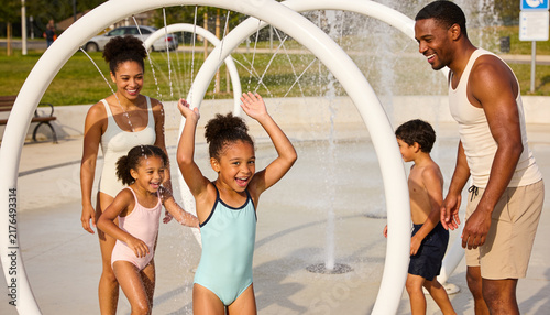 Happy Black family playing at a splash pad in a park on a sunny summer day, parents and children cooling off with water jets for outdoor recreation and summer fun