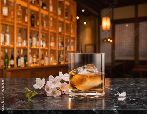 A glass of amber liquor with ice sits on a dark marble bar. Cherry blossoms adorn the counter in a traditional Japanese izakaya setting, while bottles line the shelves in the background as warm light 