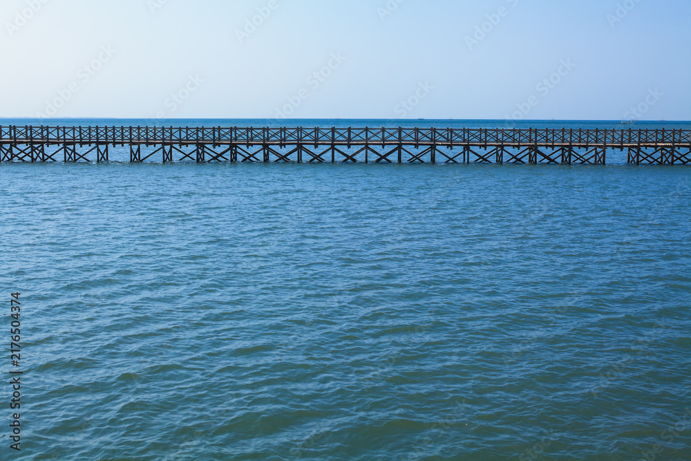 Fototapeta premium Long wooden bridge stretches across calm turquoise water. Traditional Indonesian design, serene seascape, clear blue sky create peaceful tropical atmosphere, ideal for travel and tourism promotions.
