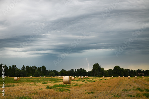 Hay bales in harvest field under dark storm clouds and distant lightning in countryside near Modena, northern Italy