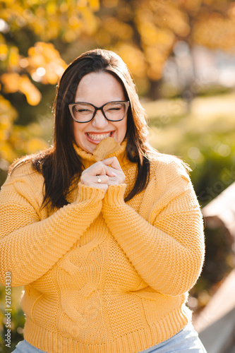 Smiling beautiful woman 30-35 year old wearing yellow knitted sweater and eye glasses holding leaf over autumn leaves close up. Fall season.