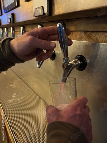 Fotografie Close up view of hands holding a glass and pulling a chrome tap handle to pour fresh pink cider or fruit beer at a self-serve tap wall in a pub