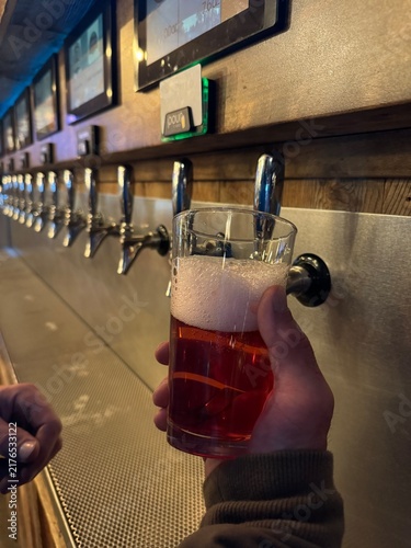 Cuadro en lienzo Person holding a pint glass filled with amber beer and foam head in front of a row of self-service draft beer taps on a stainless steel bar wall in a modern taproom