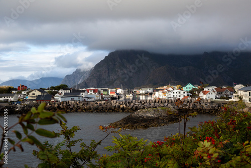 Blick auf das malerische Fischerdorf vor gigantischer Bergkulisse mit tiefen Wolken und herbstlicher Vegetation im Vordergrund, Henningsvær, Norwegen, Europa