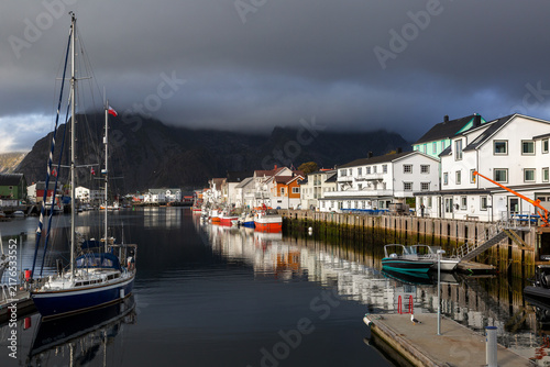 Malerischer Hafen mit Segelbooten und traditionellen Holzhäusern unter dramatischem Wolkenhimmel, Henningsvær, Norwegen, Europa