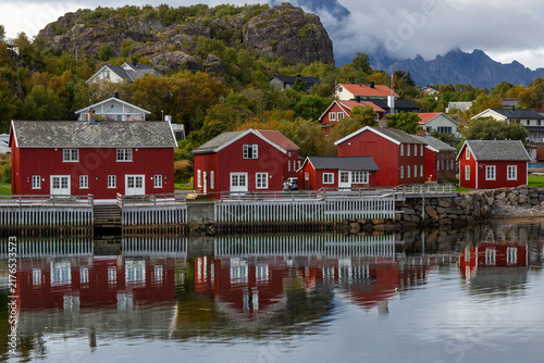 Traditionelle rote Rorbuer Fischerhütten am Wasser mit perfekter Spiegelung vor bewaldetem Hang und Bergen, Henningsvær, Norwegen, Europa