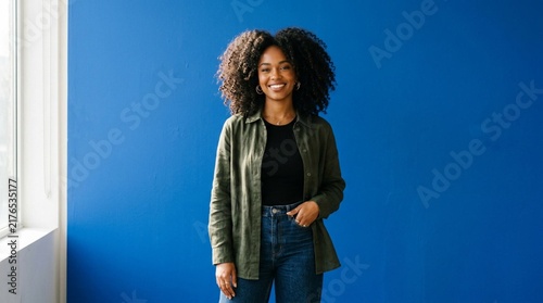 Confident young woman with voluminous curly afro hair smiling brightly against a vibrant solid blue studio backdrop, wearing a black top, olive green suede shirt, and blue denim jeans