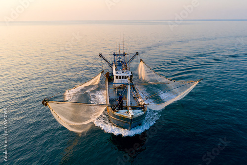 Aerial view of a commercial fishing boat trawler with large open nets moving in the blue sea water at sunset. Industrial marine fishery and seafood catch concept