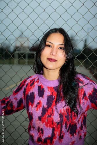 Portrait of a young confident latina woman wearing a colorful sweater outdoors by a chain link fence