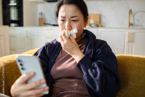 Sick woman looking at smartphone with tissue on living room sofa