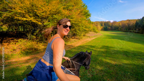Konstfotografi SELFIE: Smiling young woman riding a dark brown horse along a green meadow while colorful autumn foliage decorates the surrounding forest under a clear blue sky