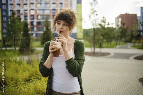Young woman stands in city park holding iced coffee. She looks thoughtful while enjoying her drink. Green trees and buildings are visible in the background during afternoon