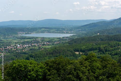 Panoramic view of a mountain lake, green valley and small village surrounded by forested hills in Lower Silesia, Poland. Concept of rural tourism, peaceful living and summer vacation in nature.