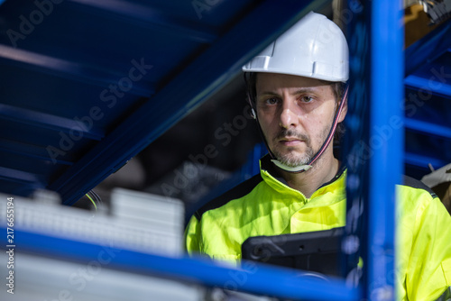 Industrial worker in hard hat and safety vest inspecting machinery inside modern factory, focused expression, blue metal structure and protective equipment