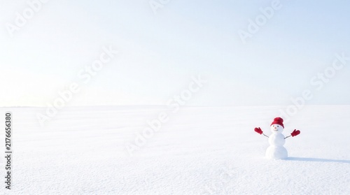 Cheerful snowman wearing a red knit hat and mittens stands in a vast white snowy field. Snowman provides copy space for winter holiday message or seasonal advertising on a minimalist bright landscape.