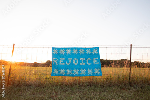 hand made quilt that says rejoice hanging from rural fence at sunset 