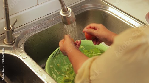 Close up of an elderly woman washing fresh red tomatoes and green vegetables in a kitchen sink, preparing a healthy organic meal. Senior lifestyle and wellness concept.