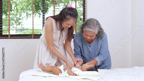 Happy multi-generational Asian family with grandmother and mother smiling while taking care and changing clothes for a cute newborn baby lying on a white bed at home.