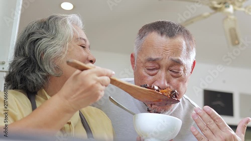 Happy senior Asian couple cooking together. An elderly husband smelling the delicious aroma of fresh homemade food from a wooden spoon held by his smiling wife in a cozy kitchen.