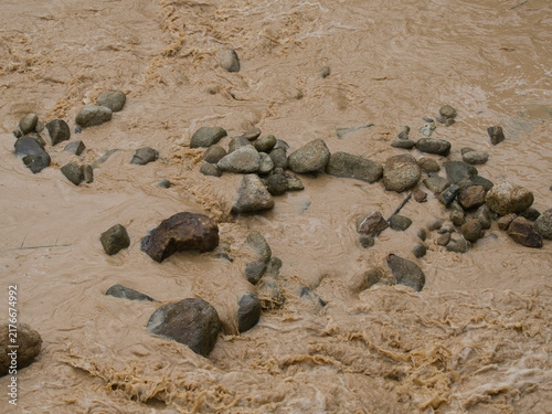 Foto Natural hazards during the rainy season river surges and mudslides rapidly flowing muddy brown water rushes past dark scattered rocks weeds and trees within the stream or river