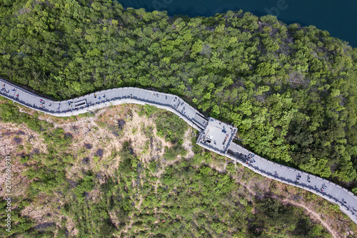 Fotografía Aerial shot of the ancient Great Wall winding over forested hills beside a body of water, with visitors walking along its stone path