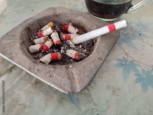Close-up of a square ashtray filled with cigarette butts and ash, with a glass cup of coffee beside it on a patterned tabletop; candid scene of a smoking ritual and morning break.