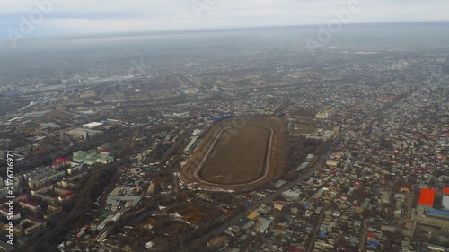 Hippodrome racetrack seen from above flying drone panning over city residential district and urban landscape under cloudy overcast sky.