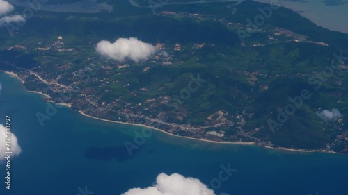 Aerial view of tropical coastline with green mountains and blue ocean beneath fluffy clouds from flight perspective.