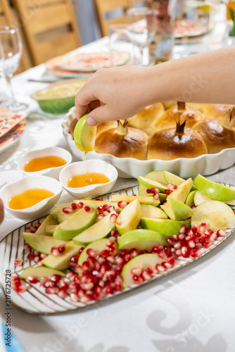 A hand dips a slice of apple into honey. A platter is filled with apple slices and pomegranate seeds. Challah bread is present on the table for Rosh Hashanah celebration.