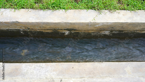 Narrow concrete drainage channel filled with flowing water beside a strip of green grass.